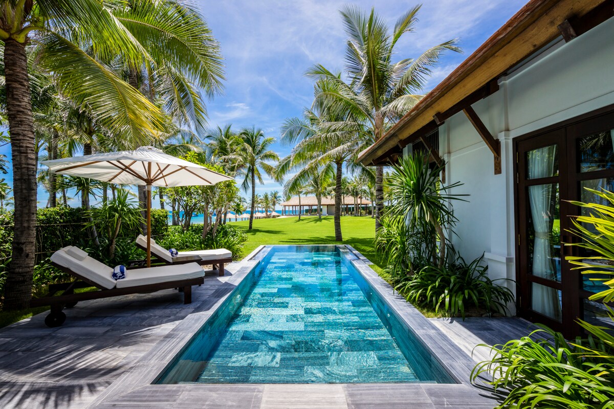 A serene private pool sits bordered by lush greenery and tropical palm trees, with sun loungers inviting relaxation nearby. The beautifully tiled pool reflects the bright sky, leading the eye toward the ocean in the distance, creating a tranquil outdoor retreat.