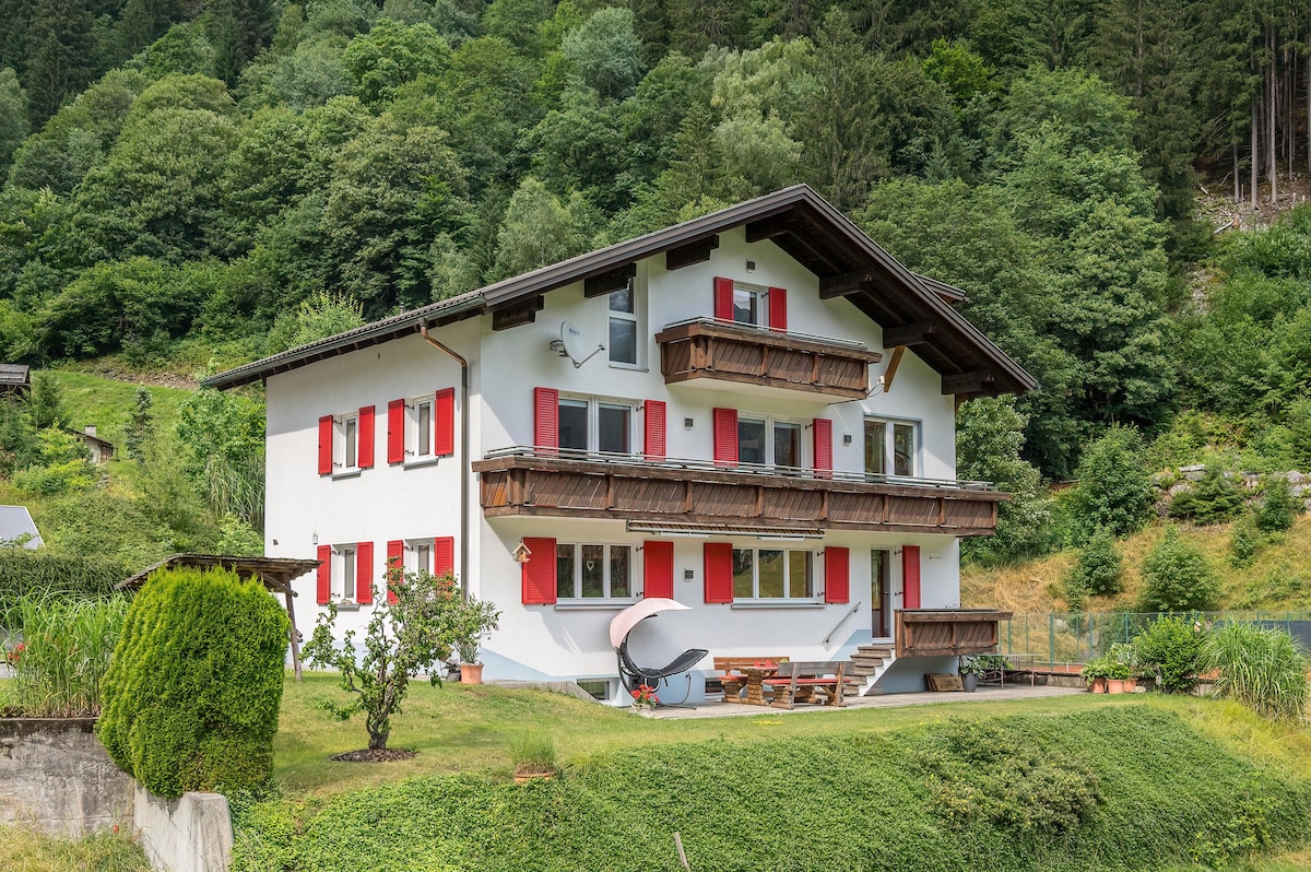 The exterior of the Alpenhaus Thurnher is shown, featuring a three-story building with white walls and red shutters. Two balconies can be seen on the upper floors, and grassy areas surround the house, complemented by trees in the background and a seating area on the ground level.