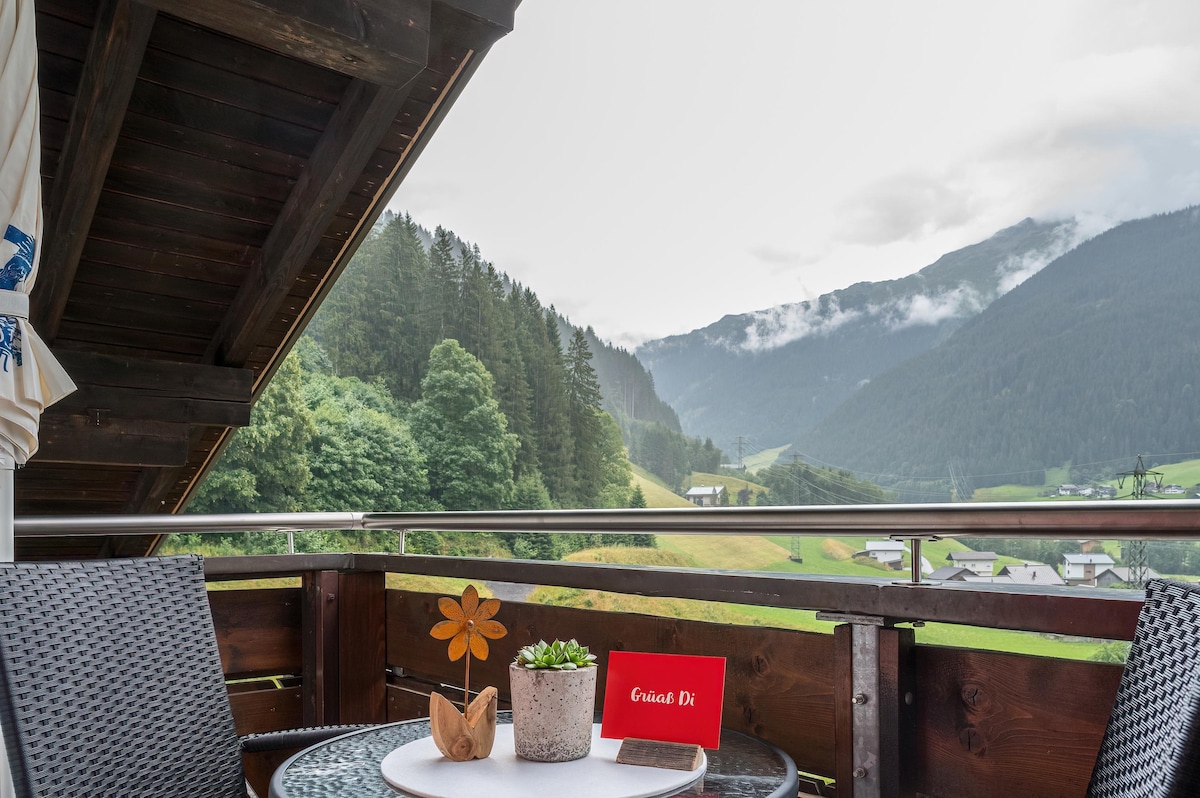 A south-facing balcony is featured, showcasing a table set with a potted flower and decorative elements. Scenic mountain views are visible in the background, framed by lush greenery, while a partly cloudy sky adds a serene ambiance to the setting.