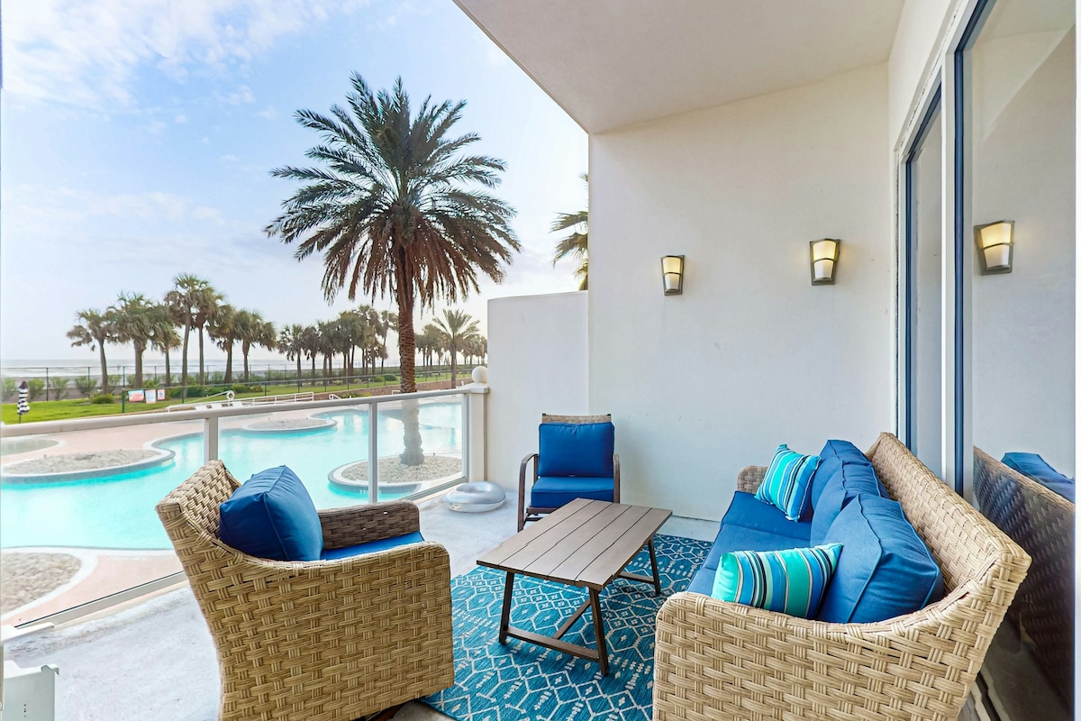 A private patio is furnished with a small table and seating for four, featuring blue cushions and decorative pillows. The setting overlooks a pool area and palm trees, with the Gulf visible in the distance. Soft natural light enhances the welcoming ambiance.