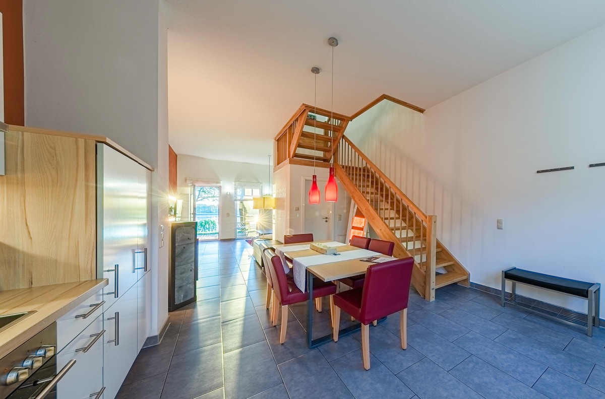 An open living area features a staircase leading to the upper level, accompanied by a dining table with red chairs. A modern kitchen is located in the background, showcasing sleek cabinetry. Large windows allow natural light to fill the space.