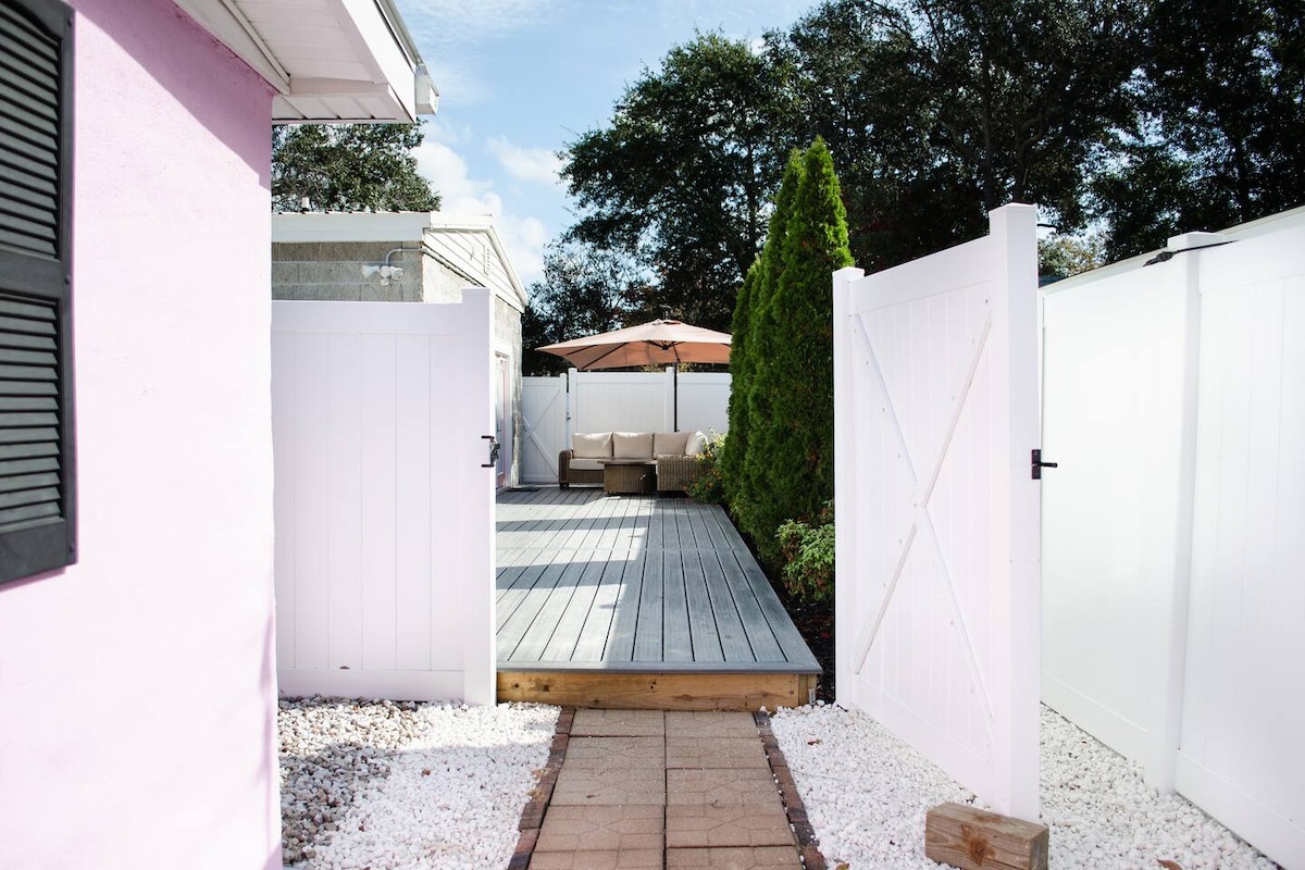 A private entrance is framed by white wooden gates leading to a spacious outdoor deck area. Comfortable seating is arranged under a large umbrella, surrounded by landscaped greenery. The path is lined with decorative stones, enhancing the inviting atmosphere of the outdoor space.