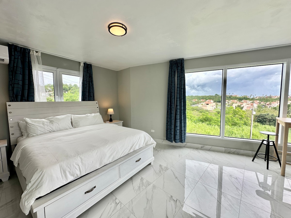 A bright and airy bedroom features a spacious bed with white linens. Large windows offer scenic views of the surrounding greenery and residential areas. Natural light illuminates the room, complemented by modern furnishings, including a bedside lamp and a small high table.