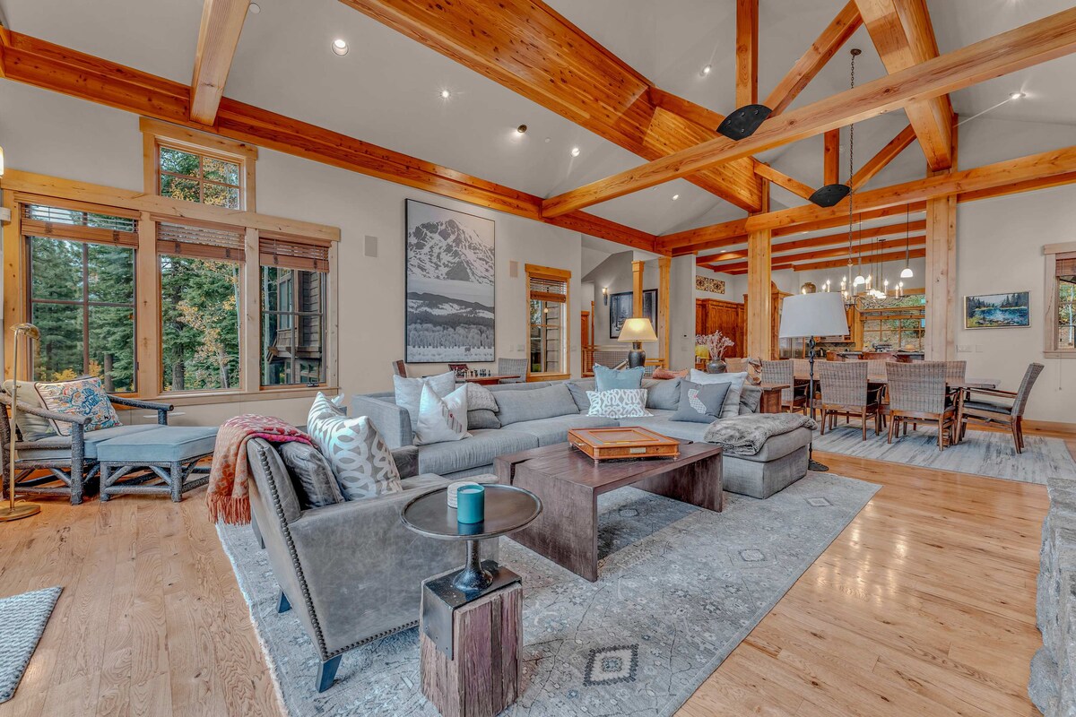 An open-concept living area is presented, highlighting a spacious sectional sofa with decorative pillows and a wooden coffee table. Exposed wooden beams accentuate the ceiling, and large windows frame views of the outdoors. A dining area with a wooden table is visible in the background.