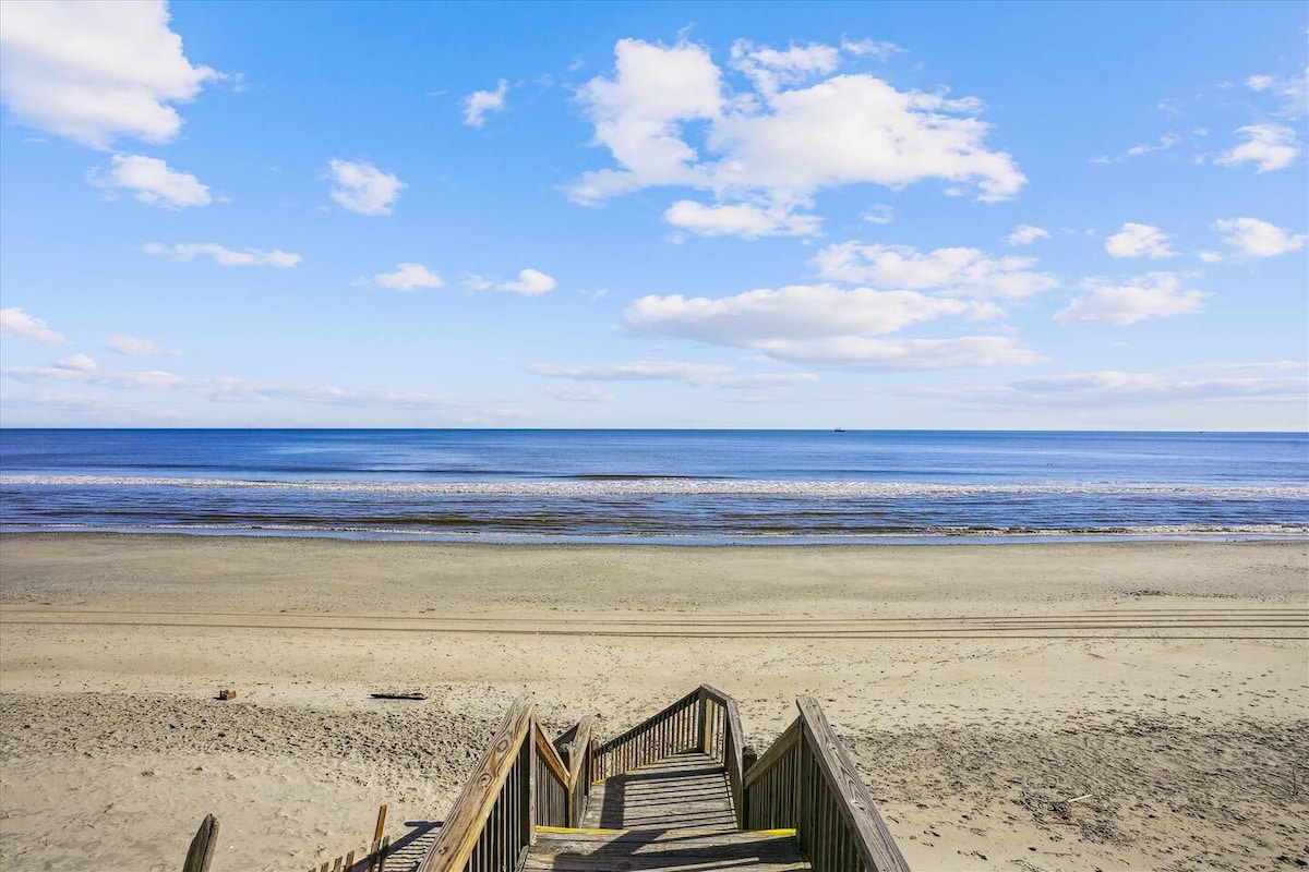 A wooden staircase leads down to the sandy beach, with gentle waves lapping at the shore. The expansive ocean stretches out under a clear blue sky dotted with a few white clouds, creating a serene coastal scene.