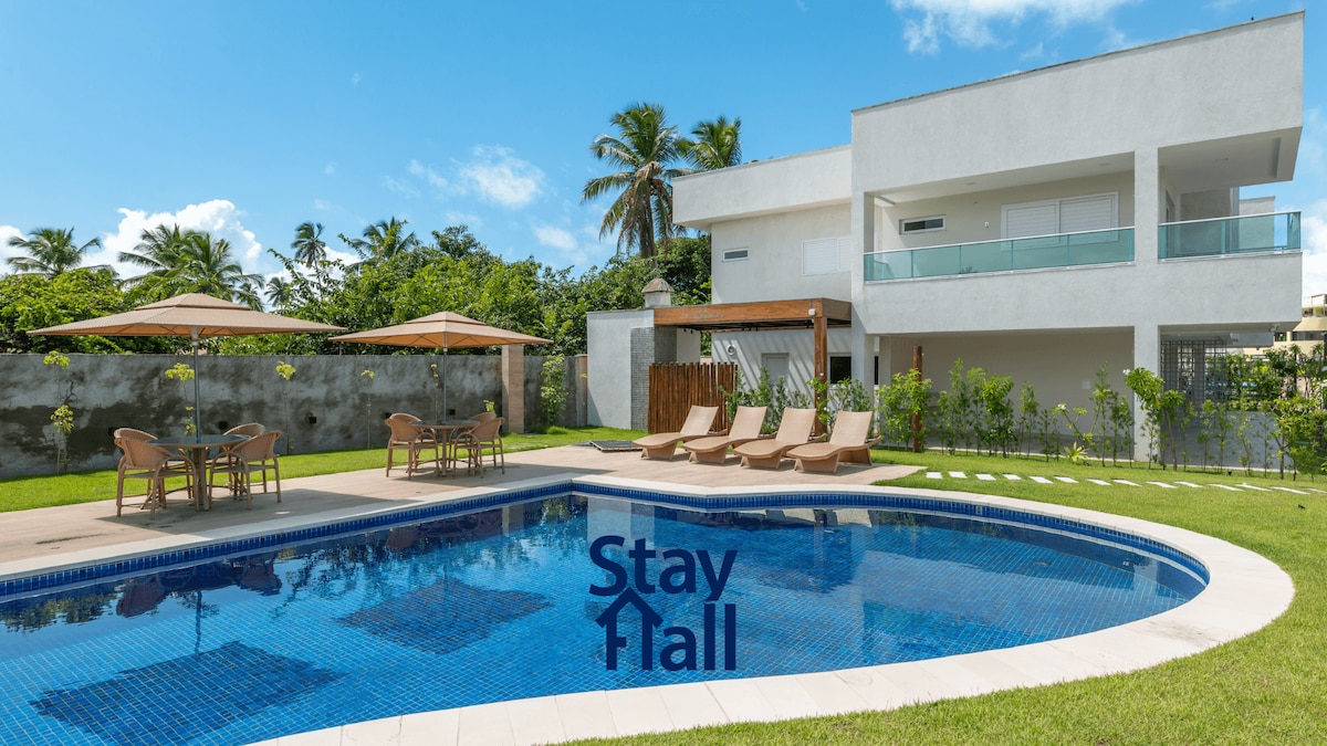 A spacious outdoor area features a large swimming pool bordered by sun loungers and shaded umbrellas. Lush greenery surrounds the property, while a two-story house displays modern architecture with a minimalistic design. A seating arrangement is positioned nearby for relaxation.
