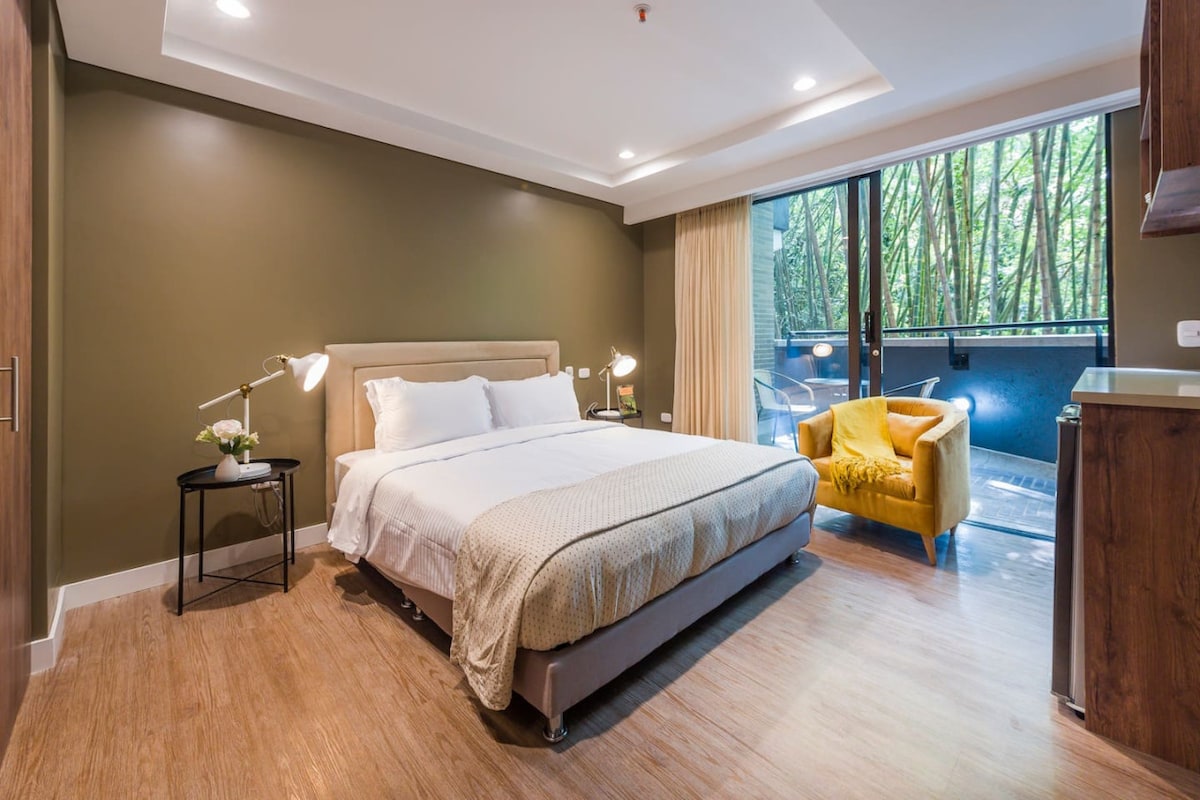 A spacious room is presented with a king-size bed covered in neutral-colored linens. A bright yellow armchair adds a pop of color, while floor-to-ceiling windows offer views of lush greenery. Soft lighting enhances the calming ambiance, and warm wooden flooring completes the look.