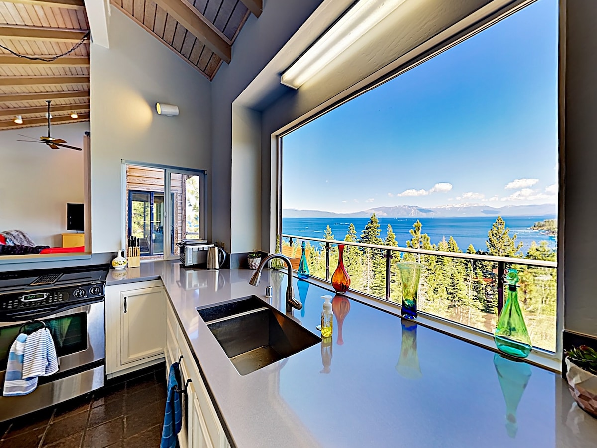 A modern kitchen features a spacious countertop and a large window showcasing panoramic views of Lake Tahoe. Colorful glass vases add a decorative touch on the ledge, while appliances and a sink are positioned for convenience. Natural light illuminates the area, enhancing the open feel.