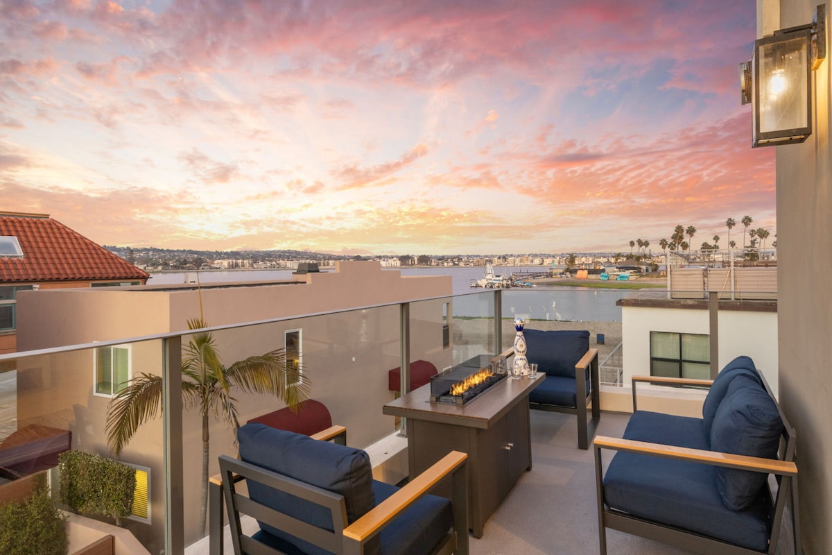 An outdoor balcony offers a serene setting with four comfortable seating options surrounding a fire pit. The bay views are complemented by a vibrant sunset, creating a stunning backdrop for relaxation.