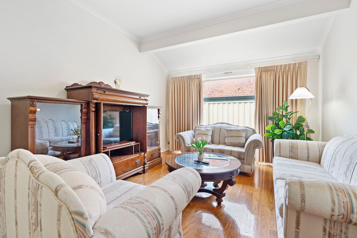 A well-lit living room is presented with hardwood flooring and large windows covered by soft curtains. Comfortable seating is arranged around a circular coffee table, complemented by a decorative plant in the corner. A wooden entertainment unit with a flat-screen TV is visible.