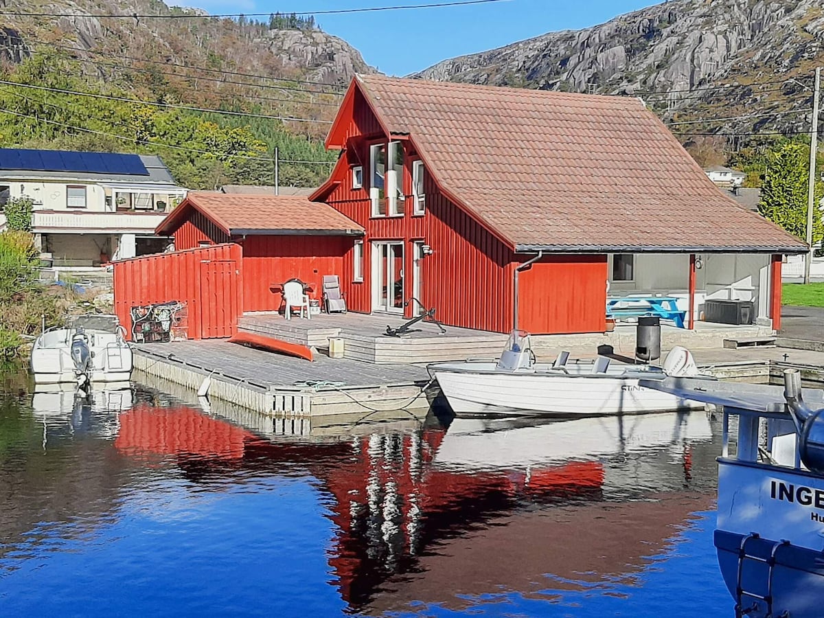 A red, modern holiday home is positioned by the water, featuring large glass windows and a spacious deck. Reflections of the house and surrounding landscape are visible on the calm water. Two boats are docked nearby, and outdoor seating is arranged on the deck.