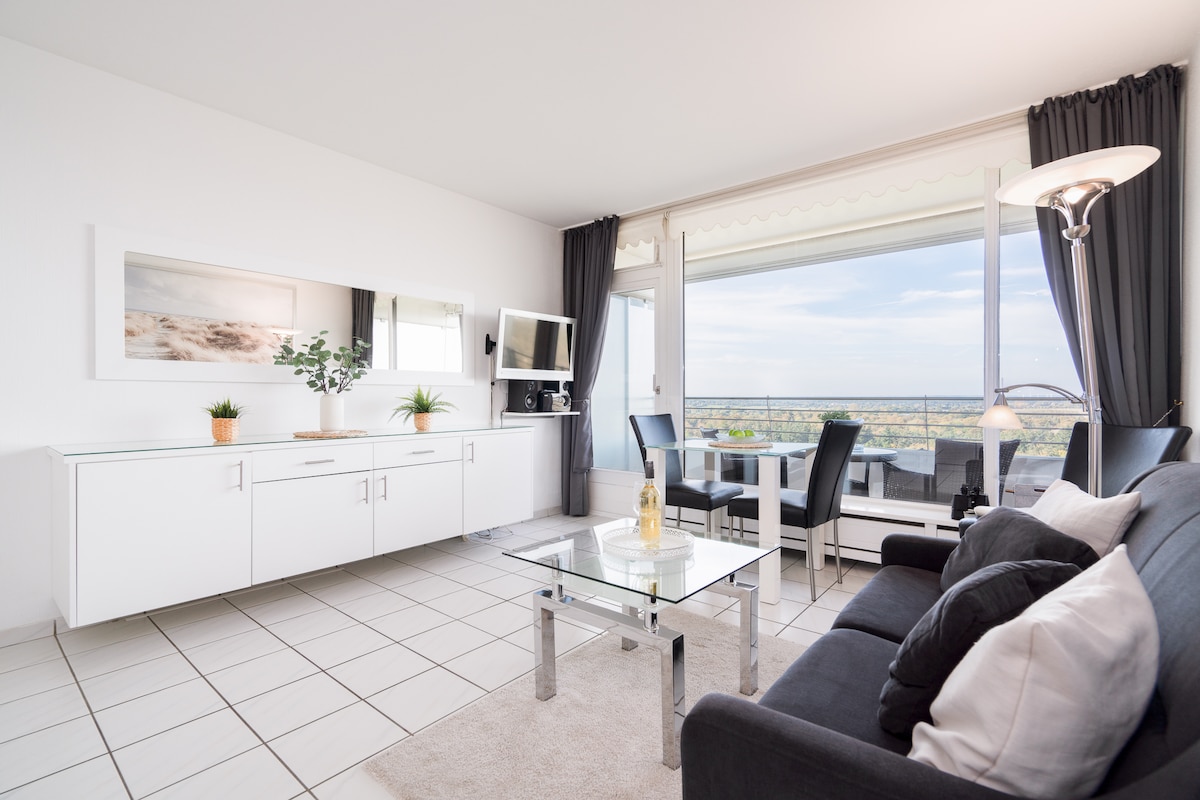 A contemporary living area features a sleek white cabinet, a flat-screen TV, and a glass coffee table. Natural light fills the space through large windows, offering views of the landscape. A dining table for four and a comfortable sofa with cushions complete the inviting ambiance.