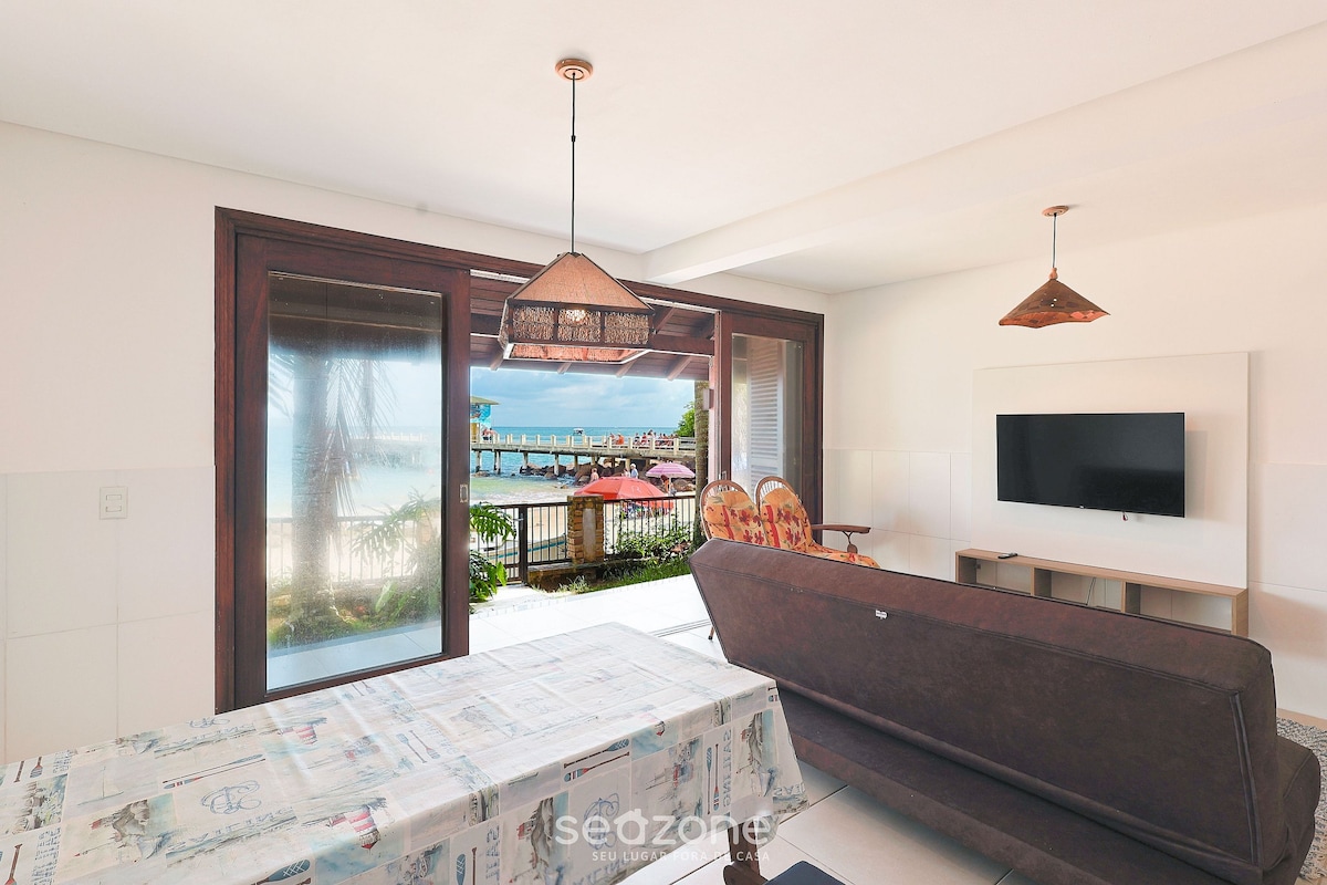 A comfortable living area is depicted, featuring a sofa facing a wall-mounted TV. Large sliding glass doors allow natural light to flood in, showcasing a view of the sea and outdoor patio. A dining table with a light tablecloth rests in the foreground.