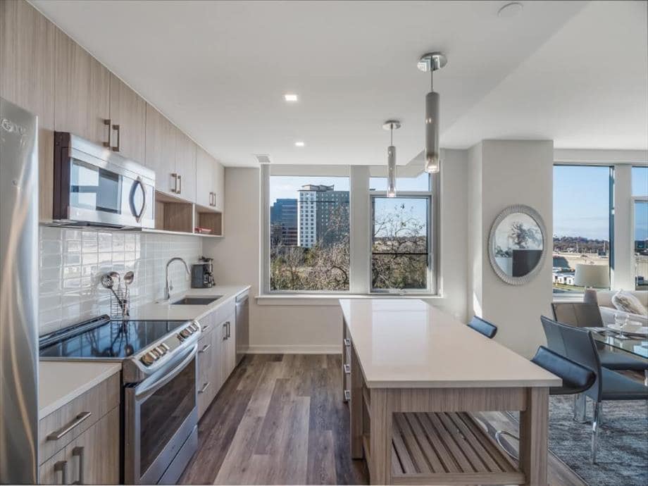 A modern kitchen is presented, featuring stainless steel appliances, including a microwave and oven. A large island with a sleek countertop offers additional preparation space. Large windows allow natural light to pour in, showcasing a view of the surrounding cityscape.