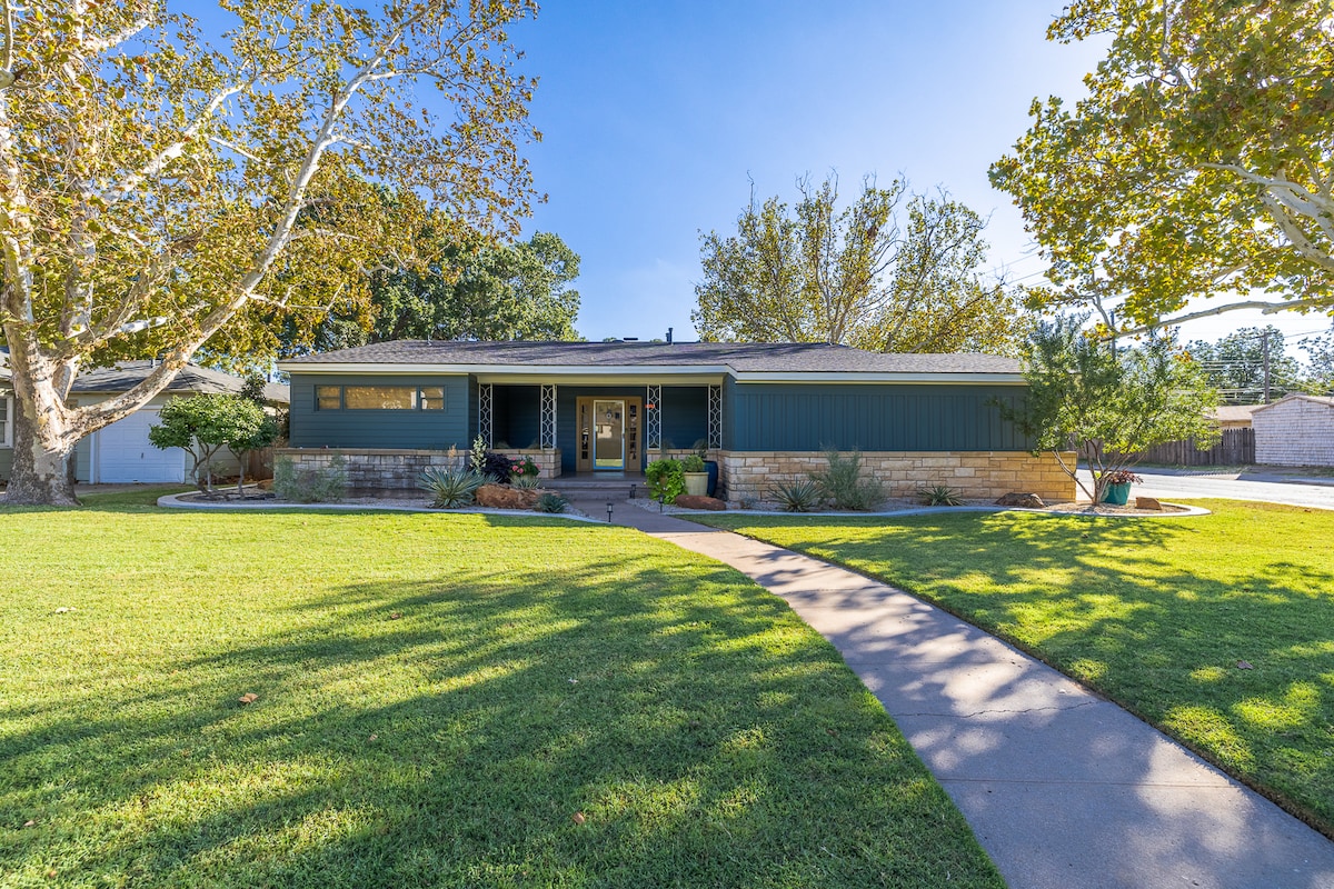 The exterior of the home is presented, showcasing a well-maintained front yard with green grass and mature trees. A paved pathway leads to the entrance, which is framed by an inviting porch and modern design elements. A large window provides natural light to the interior.