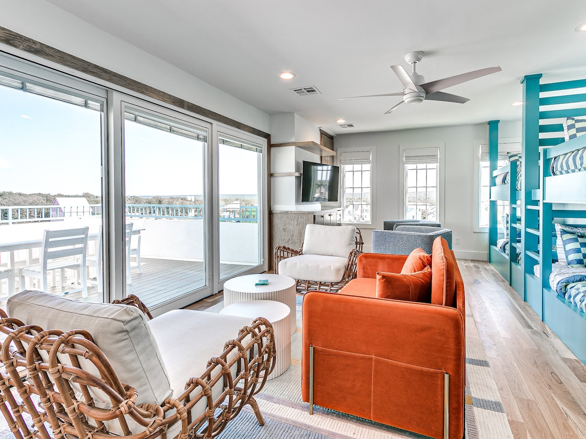 A bright and open living space is furnished with a mix of comfortable seating, including an orange sofa and light-colored armchairs. Large sliding glass doors allow for ample natural light and offer views of the outdoor area. A flat-screen TV is mounted on the wall, enhancing the leisure environment.