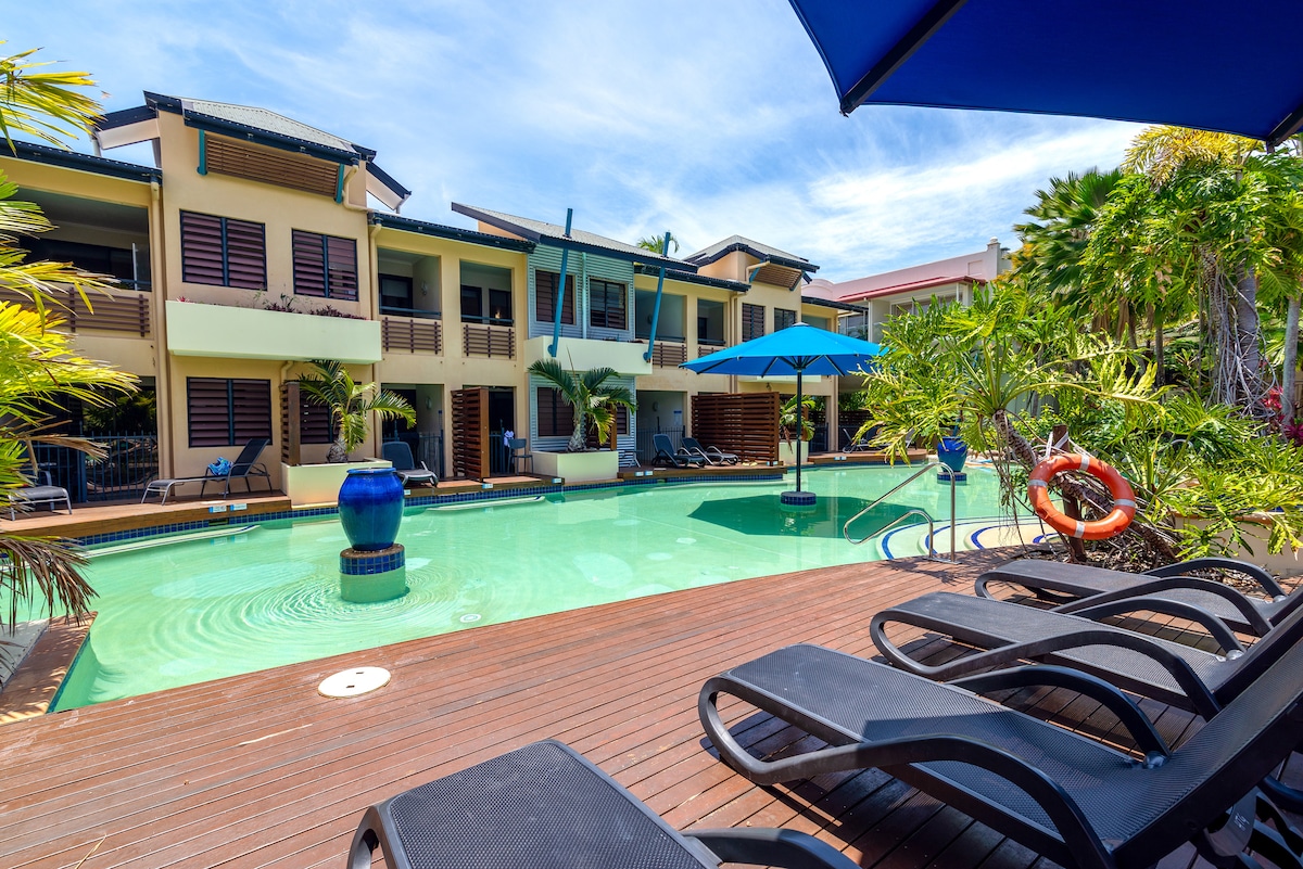 A lagoon-style pool is surrounded by palm trees and tropical foliage. Blue umbrellas provide shade over lounge chairs placed on a wooden deck. The multi-story building features balconies overlooking the pool and is complemented by a decorative blue planter.