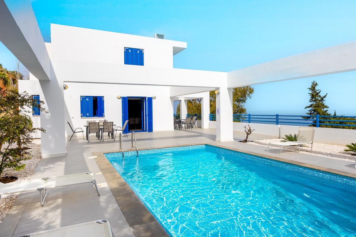 The exterior of the villa is characterized by a modern architectural design featuring white walls and blue window shutters. A private swimming pool is visible, surrounded by a paved terrace with outdoor dining furniture, offering views of the surrounding landscape.