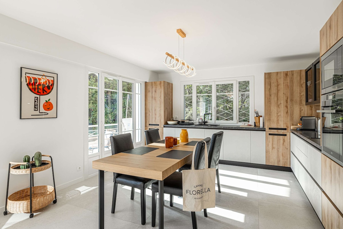 A modern kitchen is visible, featuring light cabinetry and warm wooden accents. A dining table is set with dark chairs, and multiple windows provide ample natural light. The countertop is equipped with amenities, including a black microwave and a kettle. A stylish light fixture hangs above the table.