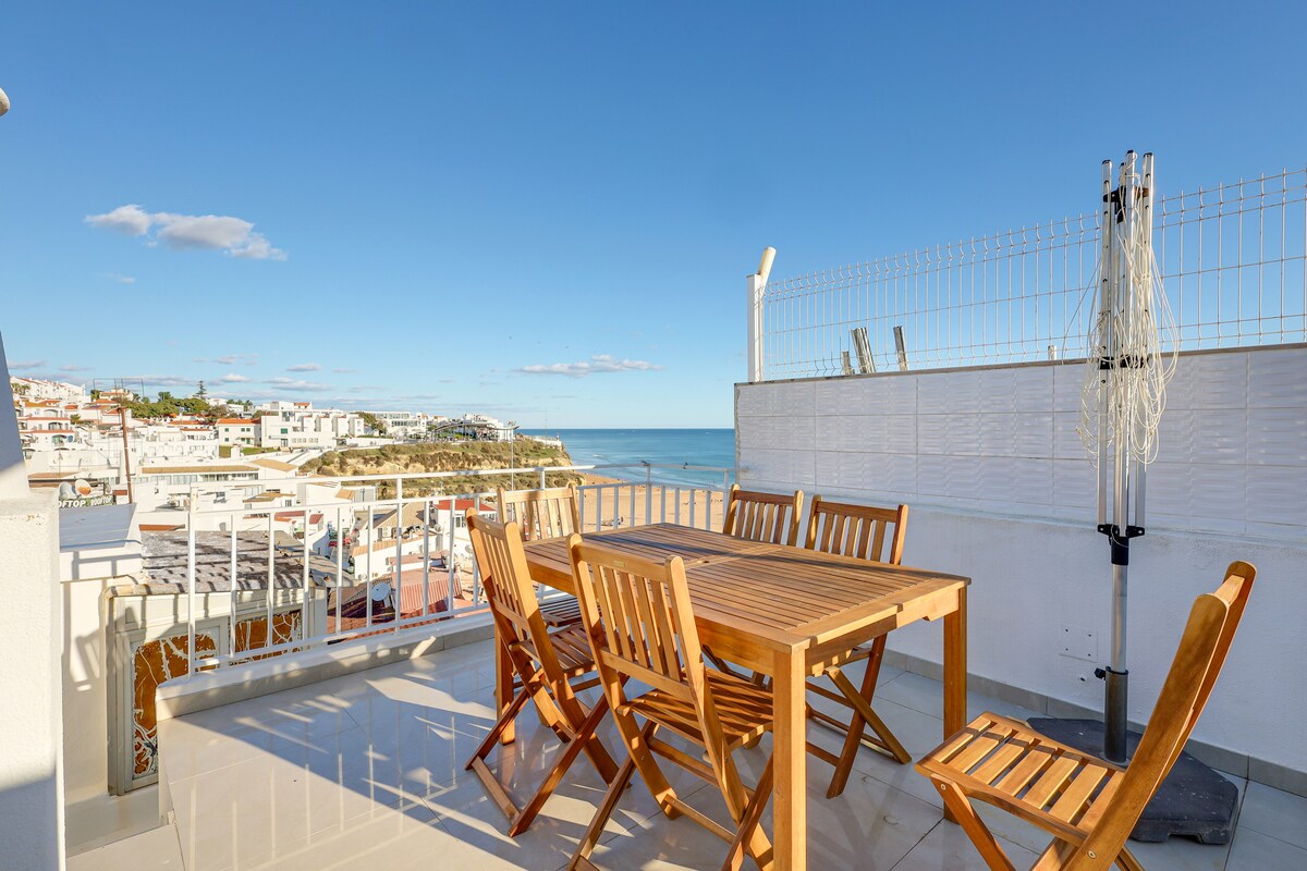 A spacious rooftop terrace features a wooden dining table accompanied by four matching chairs, all set against a backdrop of the sea and the scenic coastline. The area is bathed in natural light, with clear blue skies enhancing the serene atmosphere.
