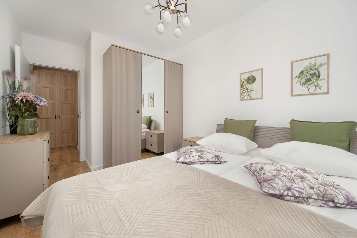  Një dhomë gjumi e rehatshme me një krevat të zbukuruar me jastëkë jeshilë dhe ngjyrë bezhë. Perdet në sfond i japin dhomës një pamje elegante.