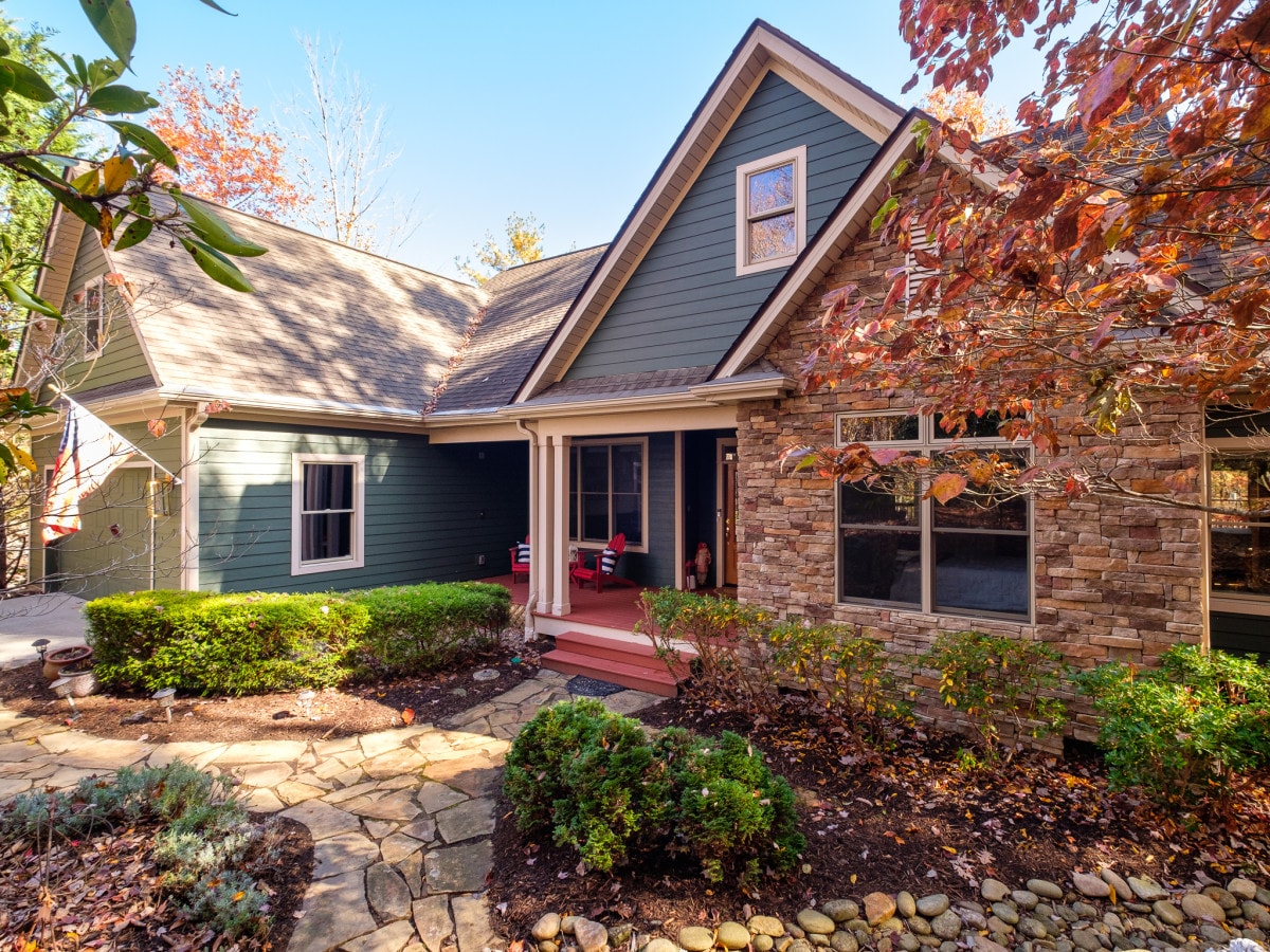 A charming exterior view of the house is highlighted by natural stone accents and a green facade. Seasonal foliage surrounds the home, with well-maintained landscaping featuring stone pathways and manicured bushes. Large windows invite natural light, creating a welcoming entrance.