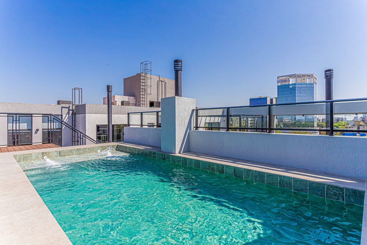 A rooftop pool is surrounded by modern railings, offering views of the cityscape. Sunlight reflects off the crystal-clear water, enhancing the serene atmosphere. Lounge areas are visible nearby, inviting relaxation under the open sky.