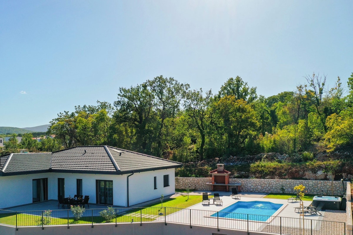 The villa, situated on a spacious property, is complemented by a private heated swimming pool. Lush trees border the yard, providing a serene backdrop. The outdoor space features lounge chairs for relaxation and a stone grill area, perfect for outdoor dining.