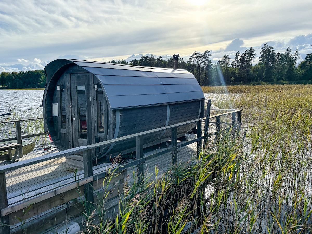 A uniquely designed sauna is situated on a wooden dock, overlooking a serene lake. Tall grasses gently sway in the breeze, and trees are visible in the distance. Soft sunlight casts a warm glow on the structure, enhancing the tranquil setting.