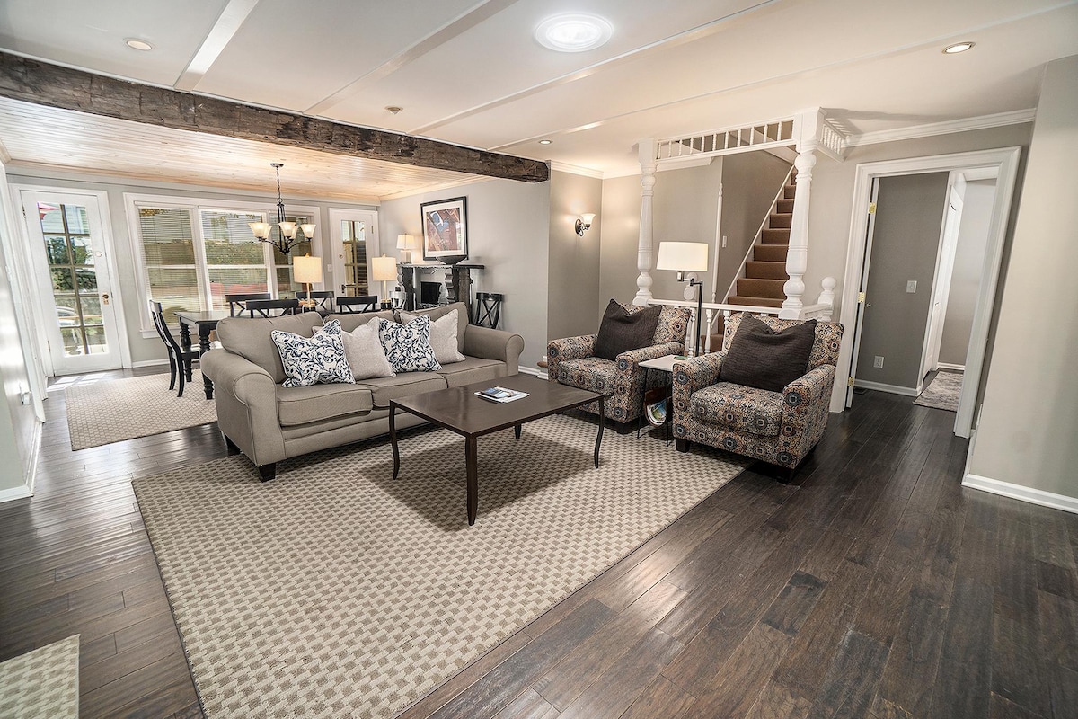 A spacious living area is featured, showcasing comfortable seating arrangements with intricately patterned chairs and a neutral sofa. A dining area is visible in the background, illuminated by natural light from large windows. Wooden beams add character to the room, enhancing the warm ambiance.