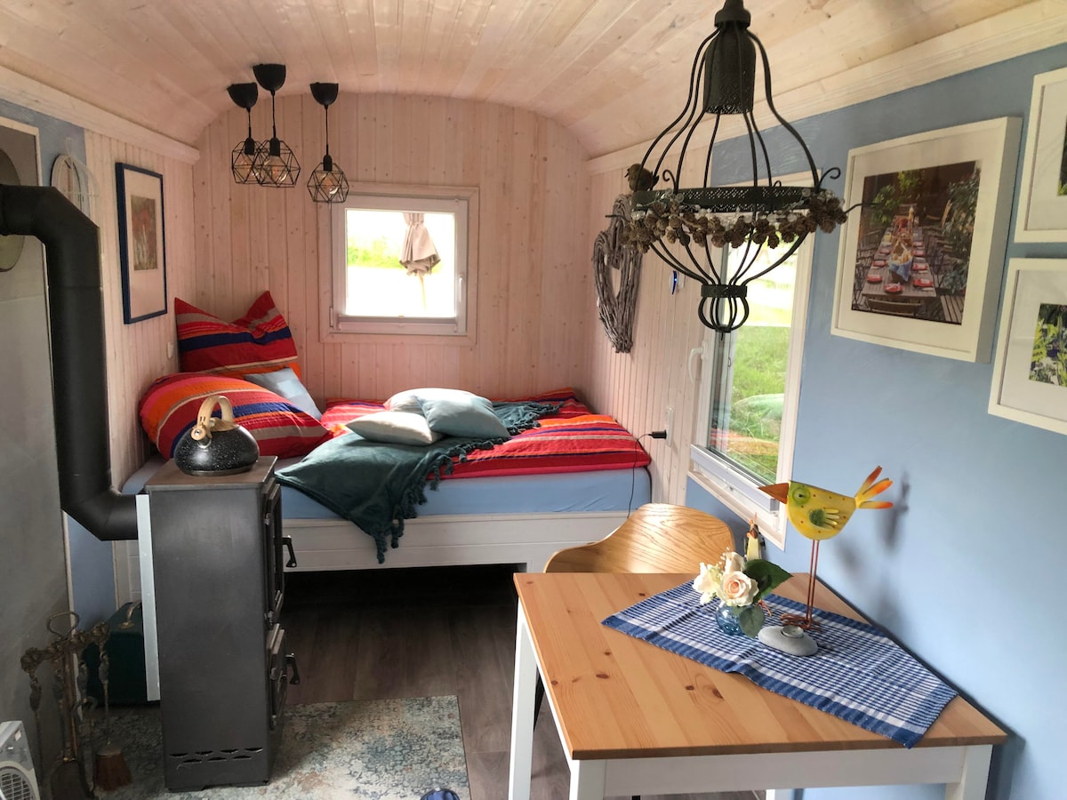The interior of the restored shepherd's hut features cozy wood paneling and brightly colored bedding on a spacious bed. A small dining table is positioned beside a window, and a decorative chandelier adds warmth to the space. A charming wood stove is visible, enhancing the inviting ambiance.