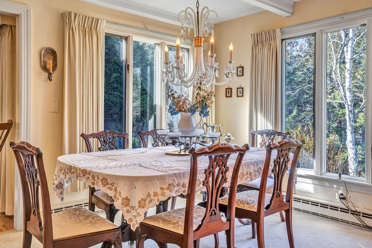 A dining area features a large table surrounded by six wooden chairs, with an elegant lace tablecloth draped across it. A chandelier hangs above, and natural light enters through three large windows framed by soft drapes, creating a warm and inviting space.
