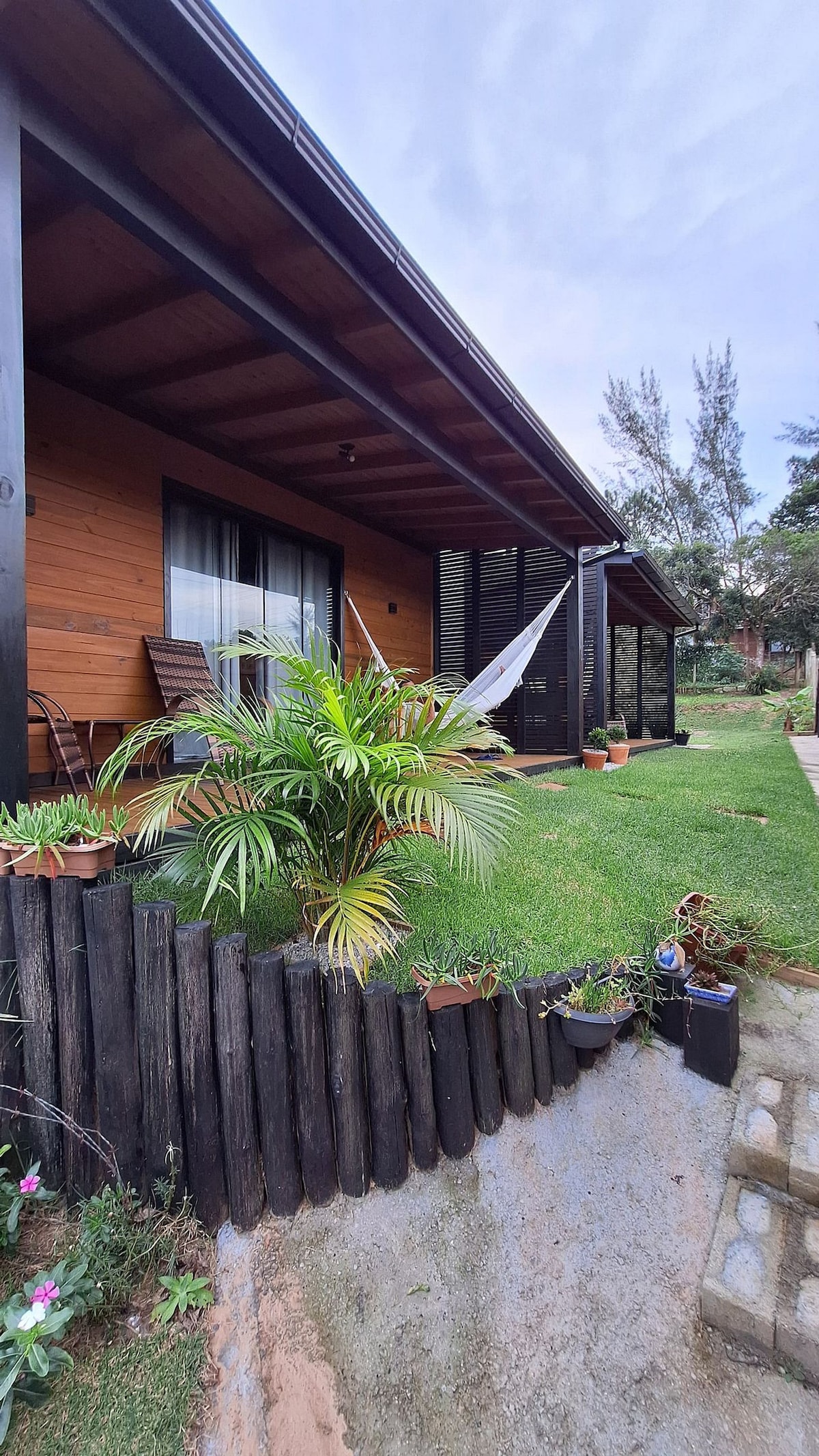 The exterior of the cabin is showcased, featuring a wooden facade and a spacious porch. A comfortable hammock is visible, surrounded by greenery and flowerpots along a low wooden fence, enhancing the natural setting of the property.
