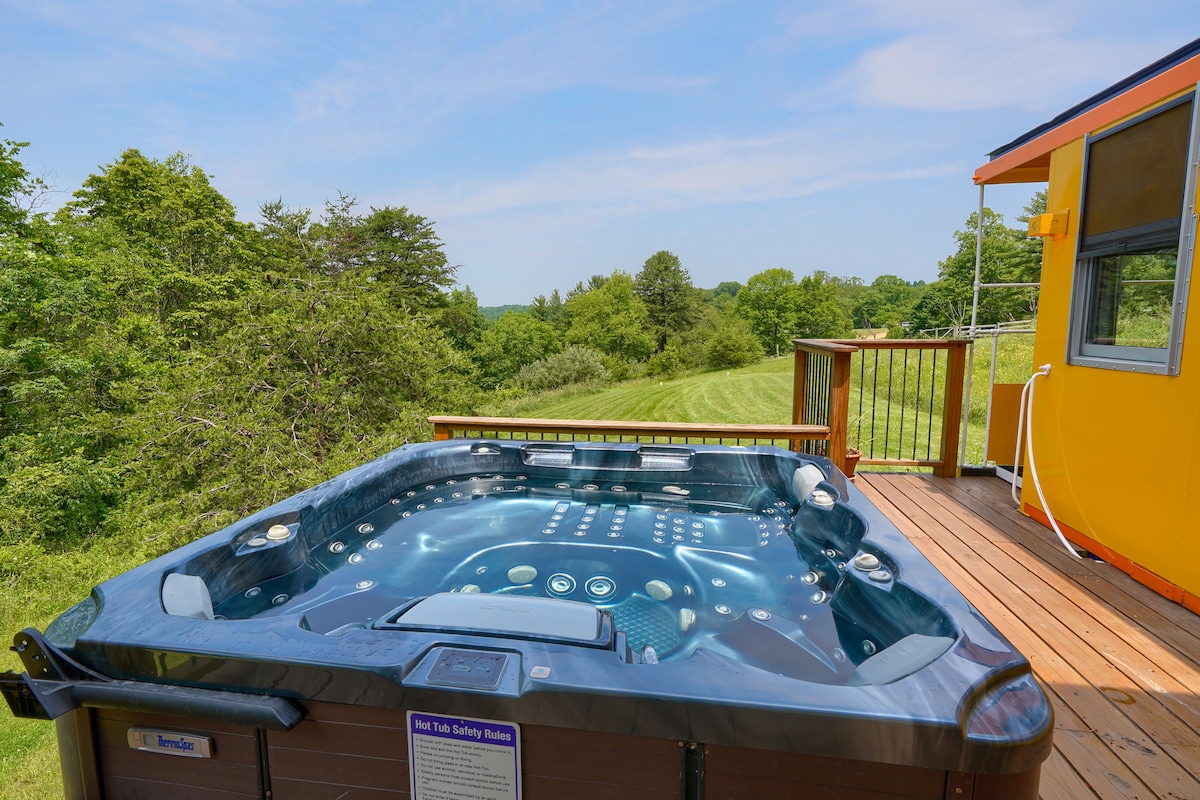 A hot tub with 126 customizable jets is positioned on a wooden deck, surrounded by lush green landscape. The clear blue sky and trees create a serene backdrop, offering a private space for relaxation and enjoyment.