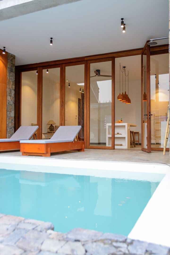 The image showcases a private pool surrounded by a stone patio. Two lounge chairs constructed of wood with light-colored cushions are positioned near the water's edge. Wide glass doors provide a view into the interior space, illuminated by pendant lighting above a dining area.