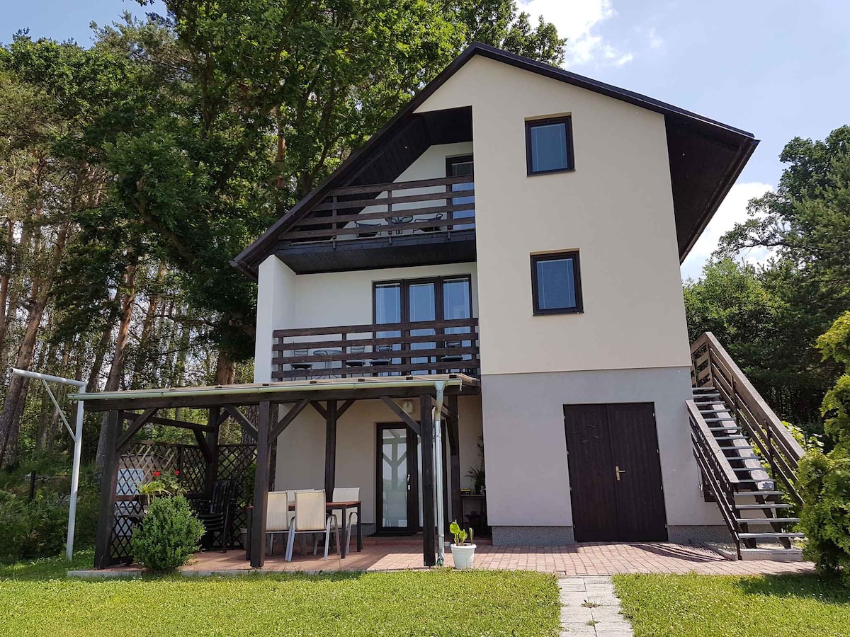 The exterior of the holiday home showcases a three-story structure surrounded by greenery. A patio area with a dining table and chairs is visible, along with outdoor steps leading to the upper floors. Large windows offer natural light, and the covered balcony adds to the charm.