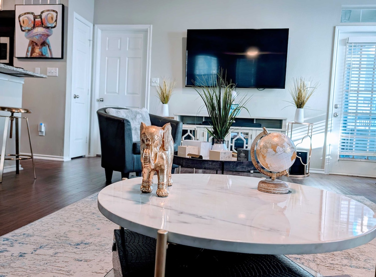 A modern living area showcases a large flat-screen TV mounted on the wall. A marble coffee table is centered, accented by decorative items including a globe and an elephant figurine. Light streams in through the sliding glass door, enhancing the inviting atmosphere.