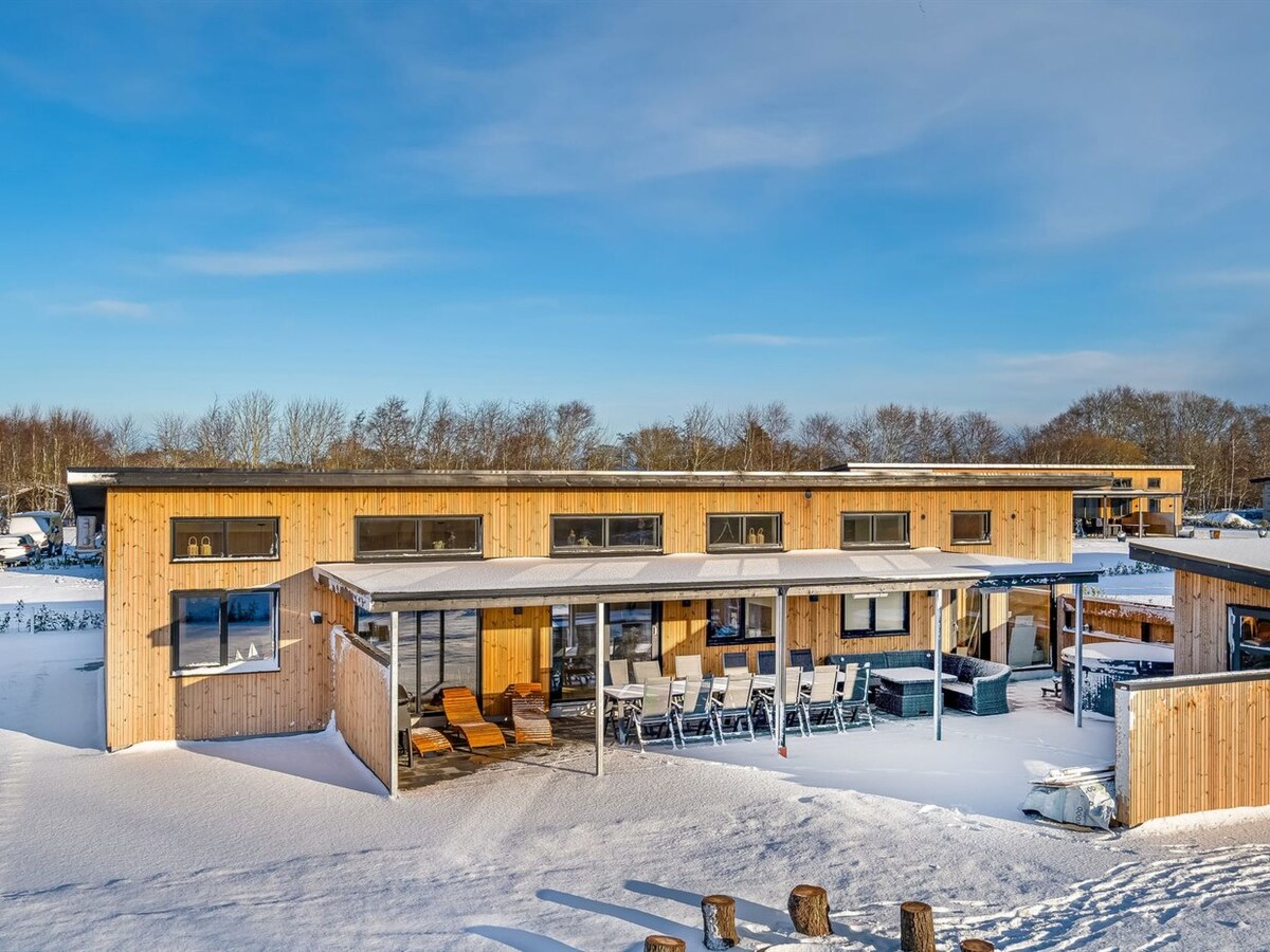 A modern two-story wooden house features a spacious exterior patio with multiple lounge chairs and a dining area, all surrounded by a snowy landscape. Large windows provide ample natural light, while a hot tub is positioned nearby for relaxation.