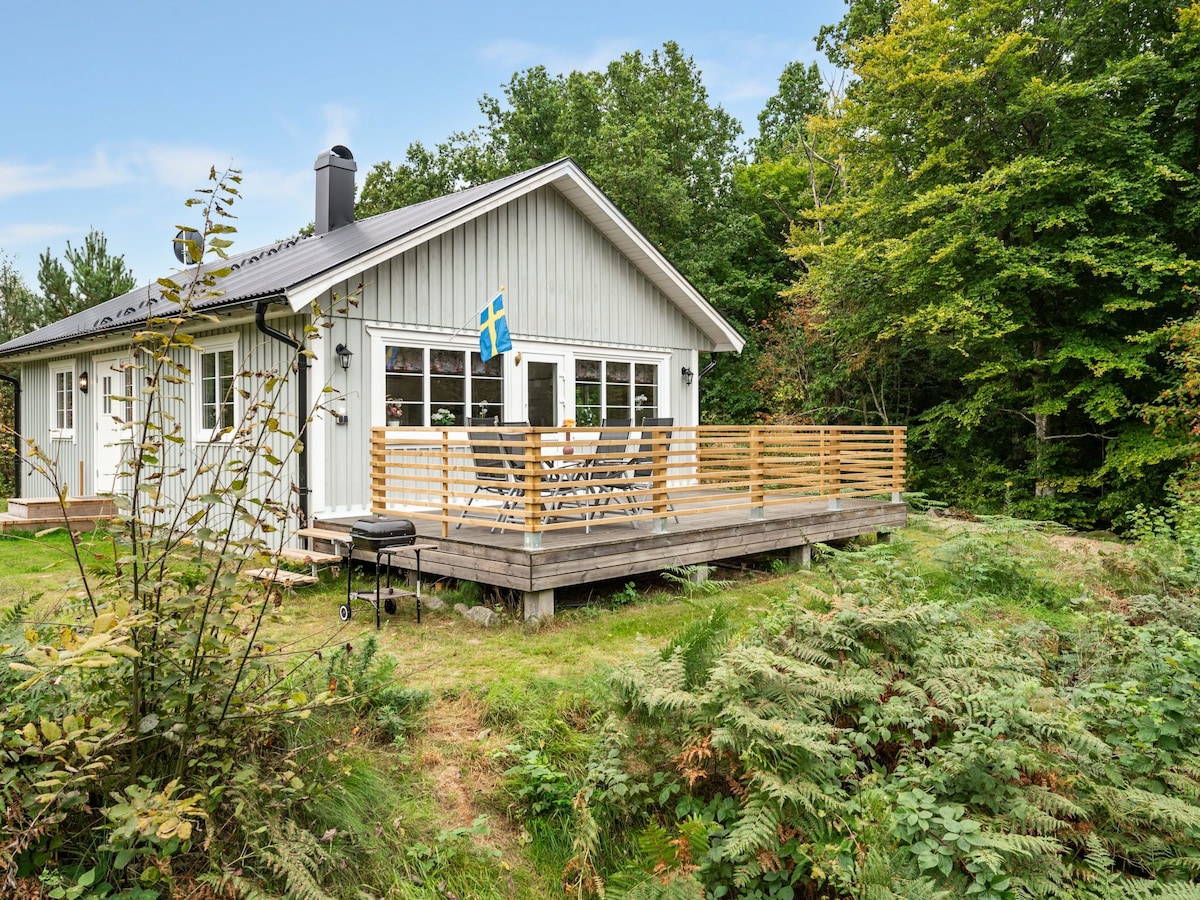 The exterior of the house is shown, featuring a wooden deck with outdoor seating and a grill. Surrounding greenery includes trees and ferns, while a Swedish flag hangs from the façade, complementing the light-colored wood of the house.
