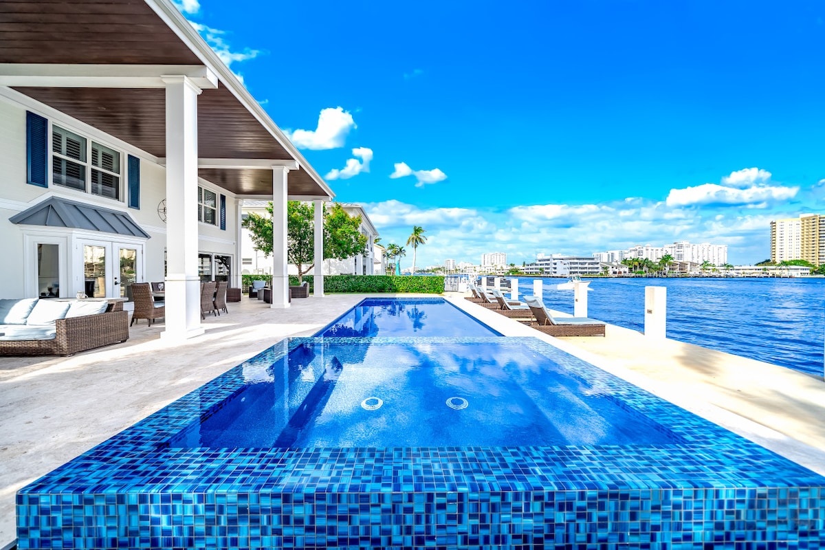 The backyard features a heated pool with a striking blue tile design, overlooking the tranquil intracoastal waterway. Lounge chairs are positioned nearby, offering a welcoming space to relax. The expansive patio area is complemented by clear blue skies and subtle greenery, enhancing the serene waterfront setting.