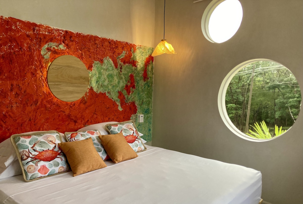 A spacious bed is adorned with decorative pillows featuring colorful patterns. A unique circular window allows natural light into the room, framing views of the greenery outside. The wall showcases a striking blend of red and green textures, complemented by a round mirror and a pendant light.
