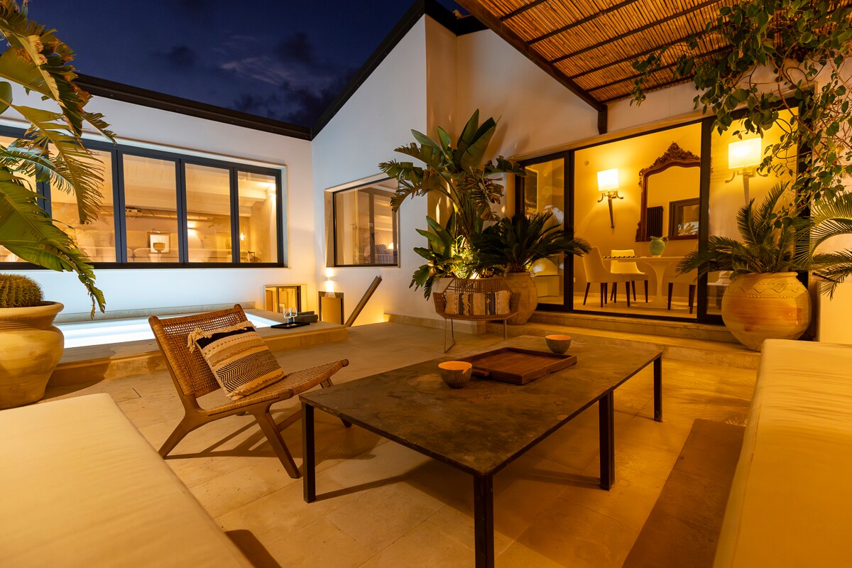 The outdoor area features a cozy seating arrangement with a low table surrounded by comfortable couches. Lush greenery adorns the space, complemented by ambient lighting from nearby lamps, creating a serene and inviting atmosphere for relaxation.