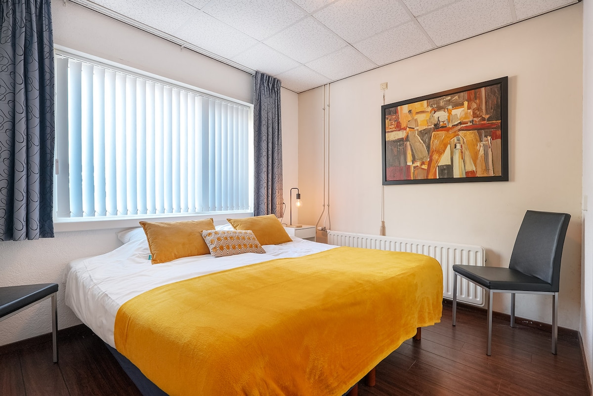A well-appointed room features a spacious bed with a bright yellow duvet and decorative pillows. Natural light enters through vertical blinds on a large window. The room includes a modern lamp and a chair, with artwork visible on the wall above the bed.