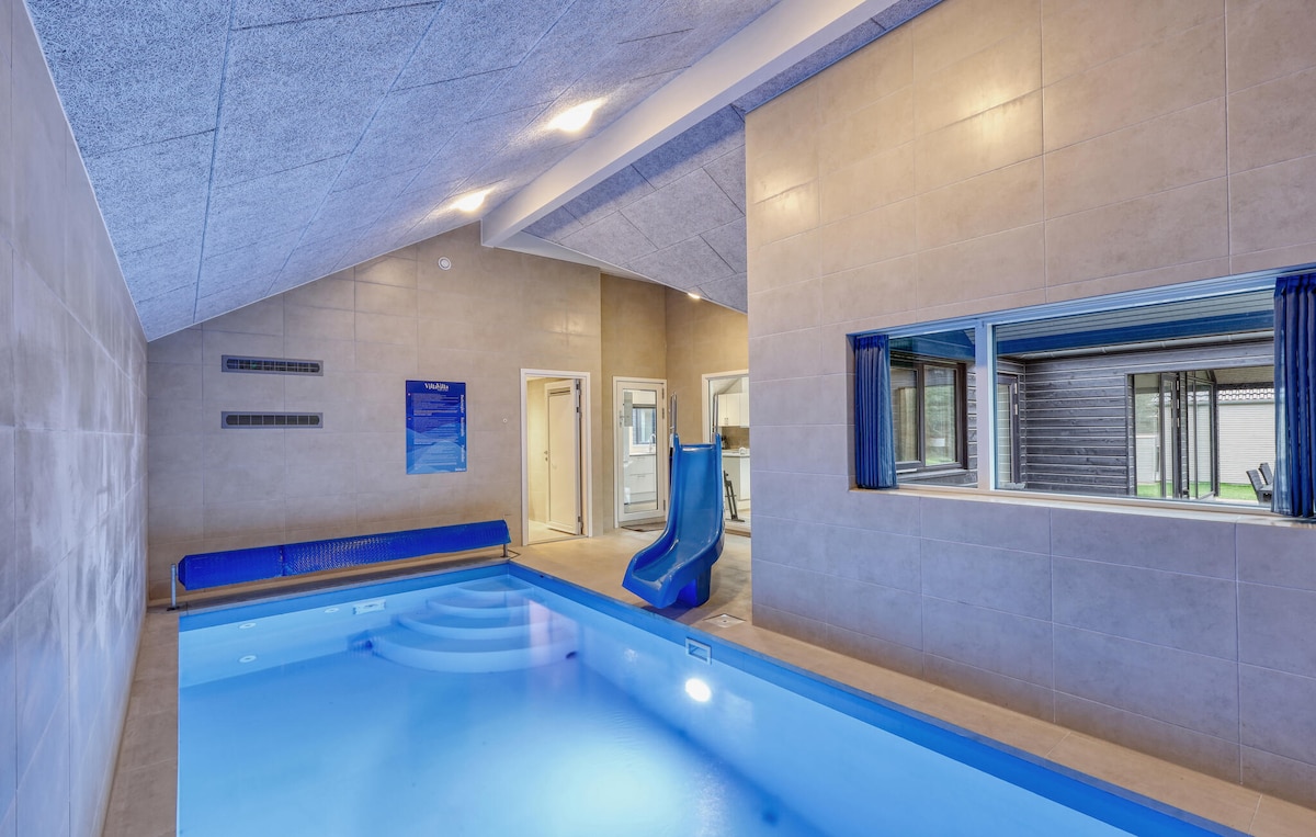 An indoor pool area features clear blue water and a curved water slide. The walls are finished with light-colored tiles, and natural light enters through a nearby window. Comfortable seating is available along one side, creating a relaxing atmosphere for guests.