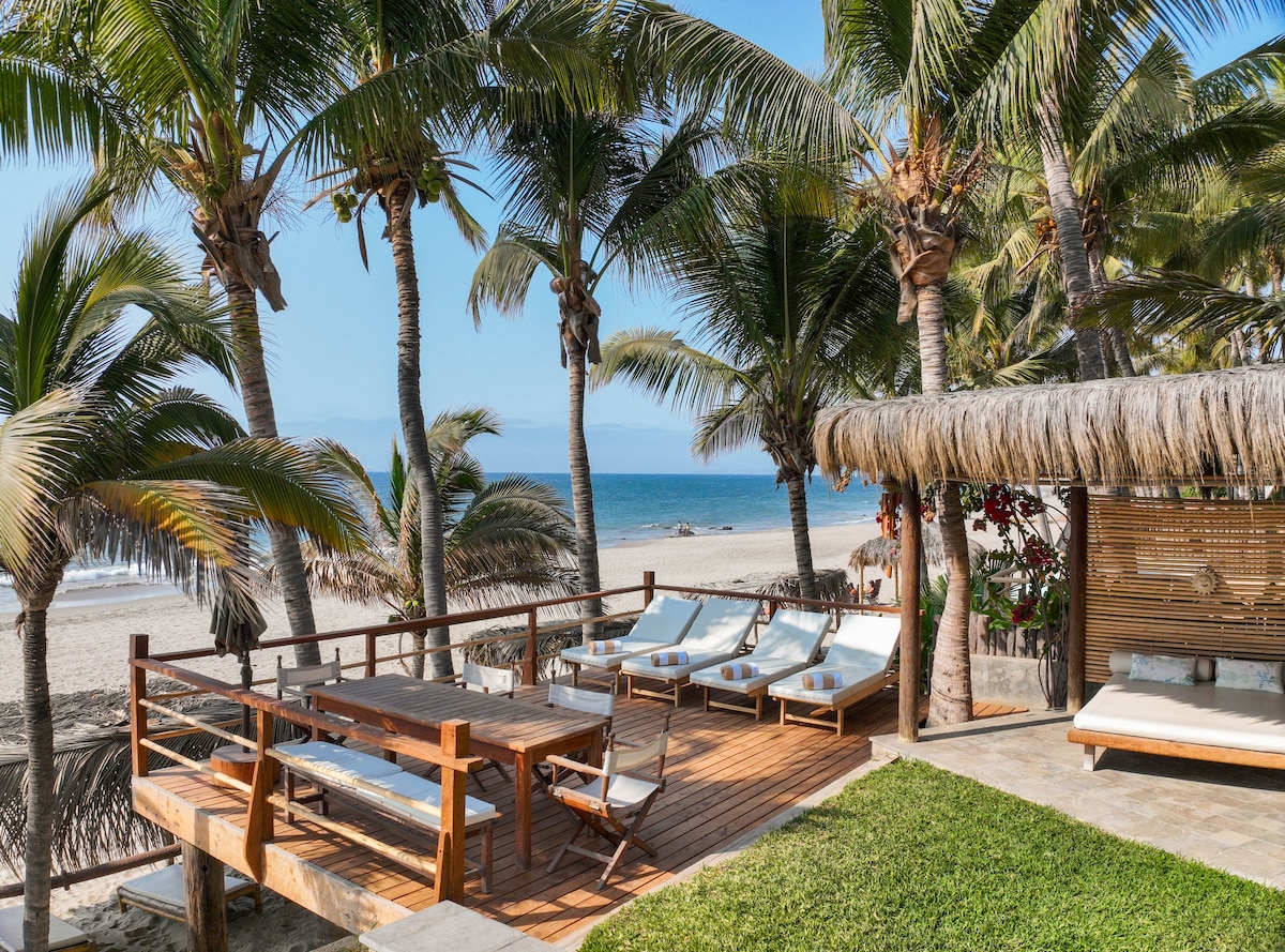 A spacious deck is surrounded by palm trees, offering a view of the sandy beach and ocean beyond. Comfortable sun loungers are positioned on the wooden surface, accompanied by a dining table and chairs, creating an inviting outdoor space for relaxation and enjoyment.