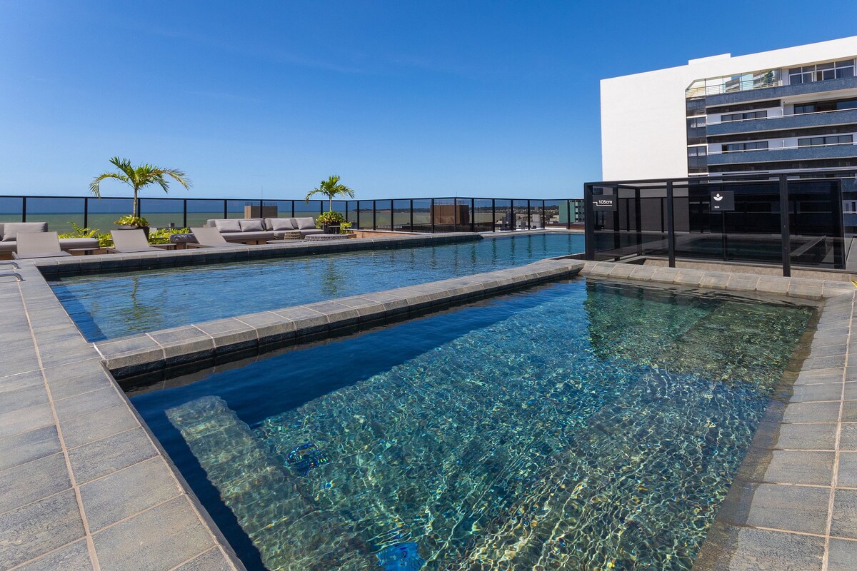Two swimming pools are visible on the rooftop, featuring clear blue water and surrounded by a sunbathing area. Deck chairs are neatly arranged along the pool edge, while tropical plants add a touch of greenery to the setting. The expansive sea view enhances the space.