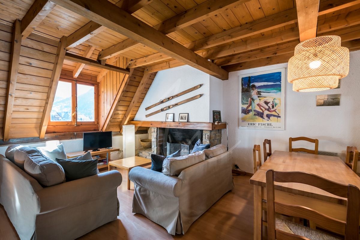 A charming living area features wooden beams and a sloped ceiling, with natural light streaming through a window. A cozy seating arrangement is positioned near a fireplace, complemented by a wooden table and chairs. A decorative light fixture adds warmth to the space.