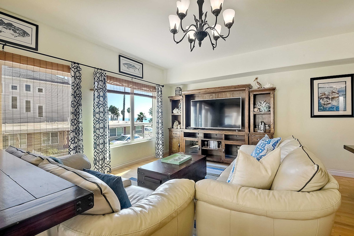 A sunlit living room is furnished with a spacious leather sofa and a wooden coffee table. A smart TV is mounted on a wooden entertainment center, with coastal-themed décor surrounding the area. Large picture windows provide views of the ocean and palm trees, accented by patterned curtains.