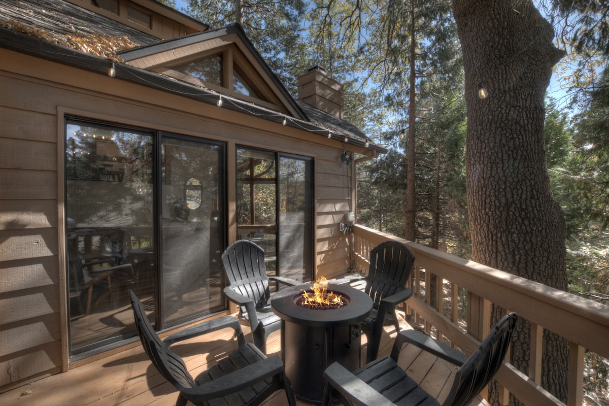 An outdoor deck is surrounded by trees, featuring a circular fire pit at the center. Four black adirondack chairs are arranged around the fire pit, providing a cozy atmosphere for gatherings. Large glass doors lead into the cabin, allowing for a seamless connection with nature.