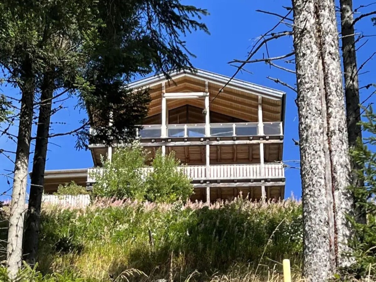 The chalet is situated on a hillside, featuring a spacious terrace with railings and a beautiful wooden façade. Surrounding greenery and wildflowers provide a natural setting, while a clear blue sky enhances the overall view of the chalet's structure.